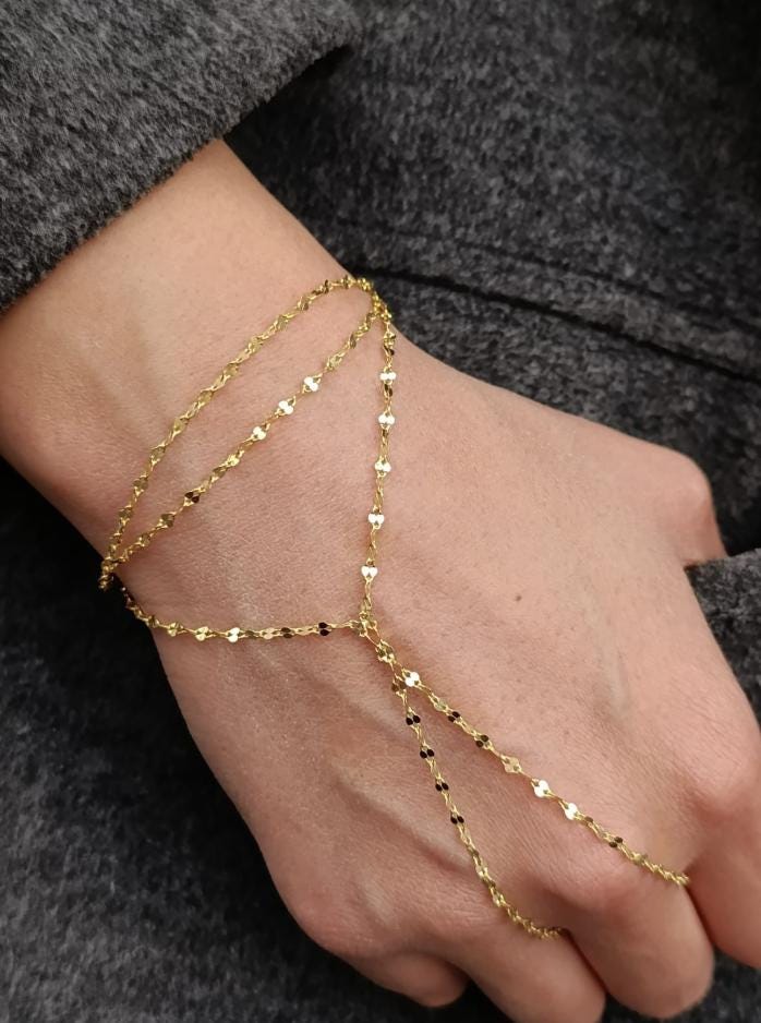 May include: A gold hand chain bracelet. The bracelet features multiple delicate chains. One chain wraps around the wrist, and another extends to connect to a ring. The chains are adorned with small, flat, circular and diamond-shaped accents. The bracelet is displayed on a hand against a gray background.