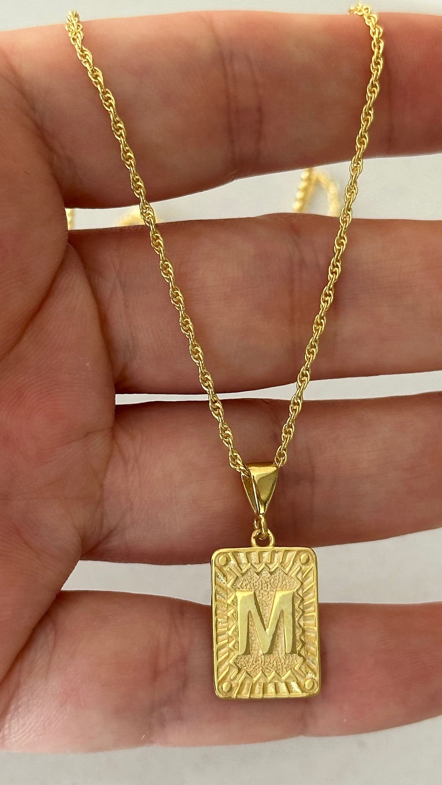 May include: A gold rope chain necklace with a gold pendant. The pendant is a square with a raised, textured border and the letter 'M' in the center.