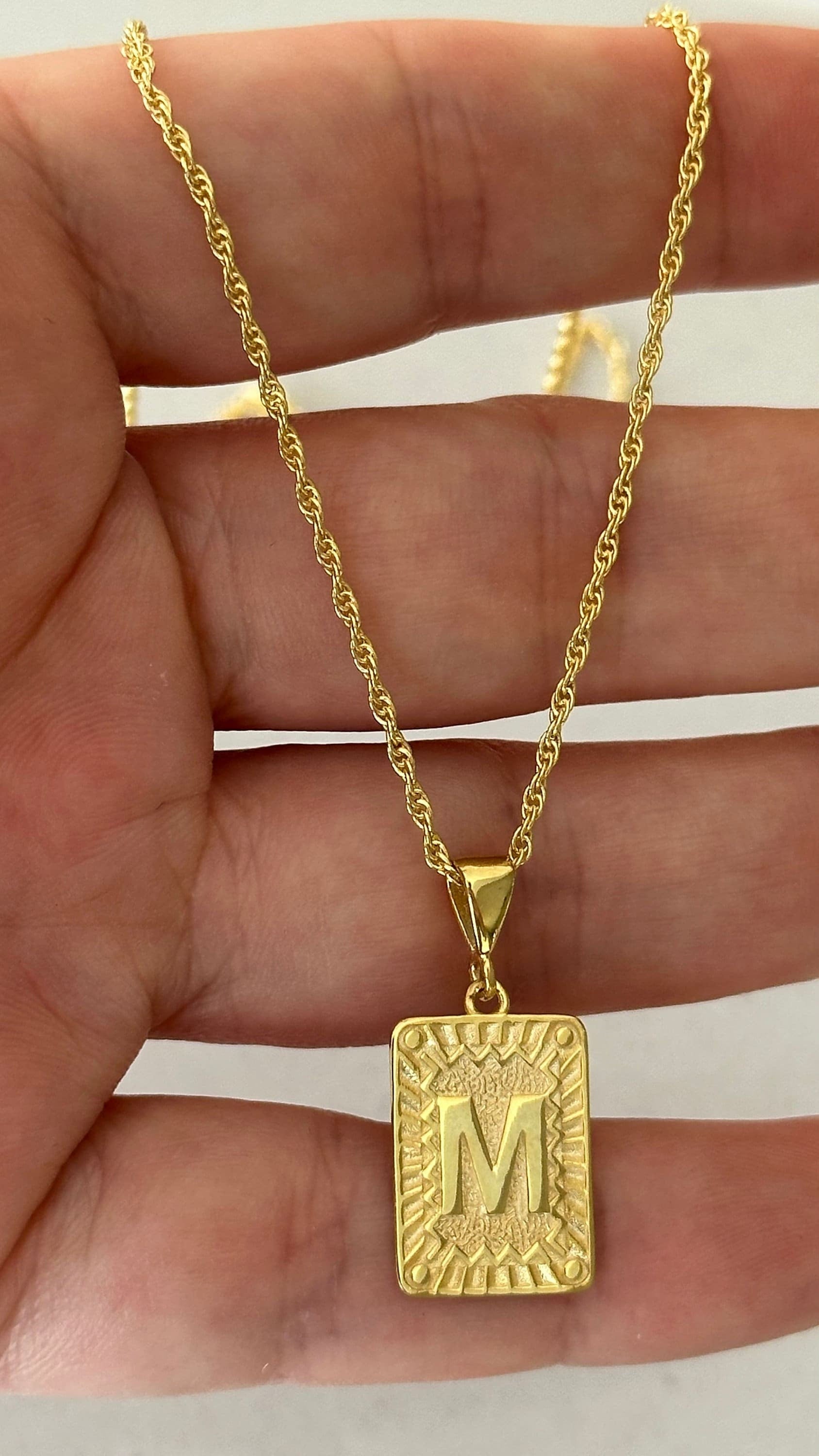 May include: A gold rope chain necklace with a gold pendant. The pendant is a square with a raised, textured border and the letter 'M' in the center.
