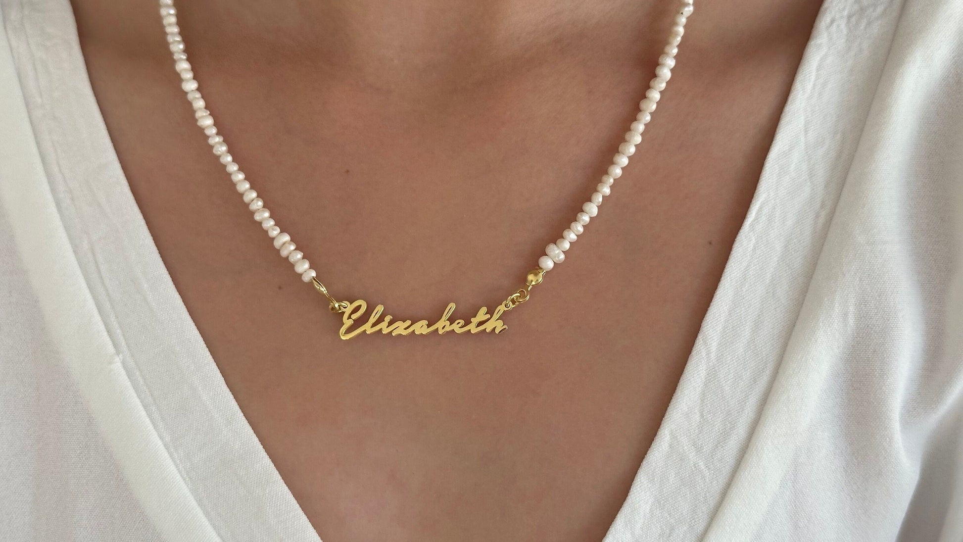 May include: A gold nameplate necklace with the name 'Elizabeth' on it. The necklace is on a white pearl chain.