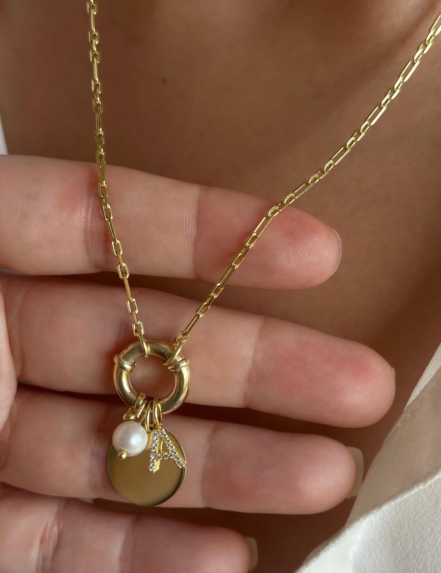 May include: A gold chain necklace with multiple charms featuring a small pearl, a diamond letter 'A', and a round disc on a toggle claps.