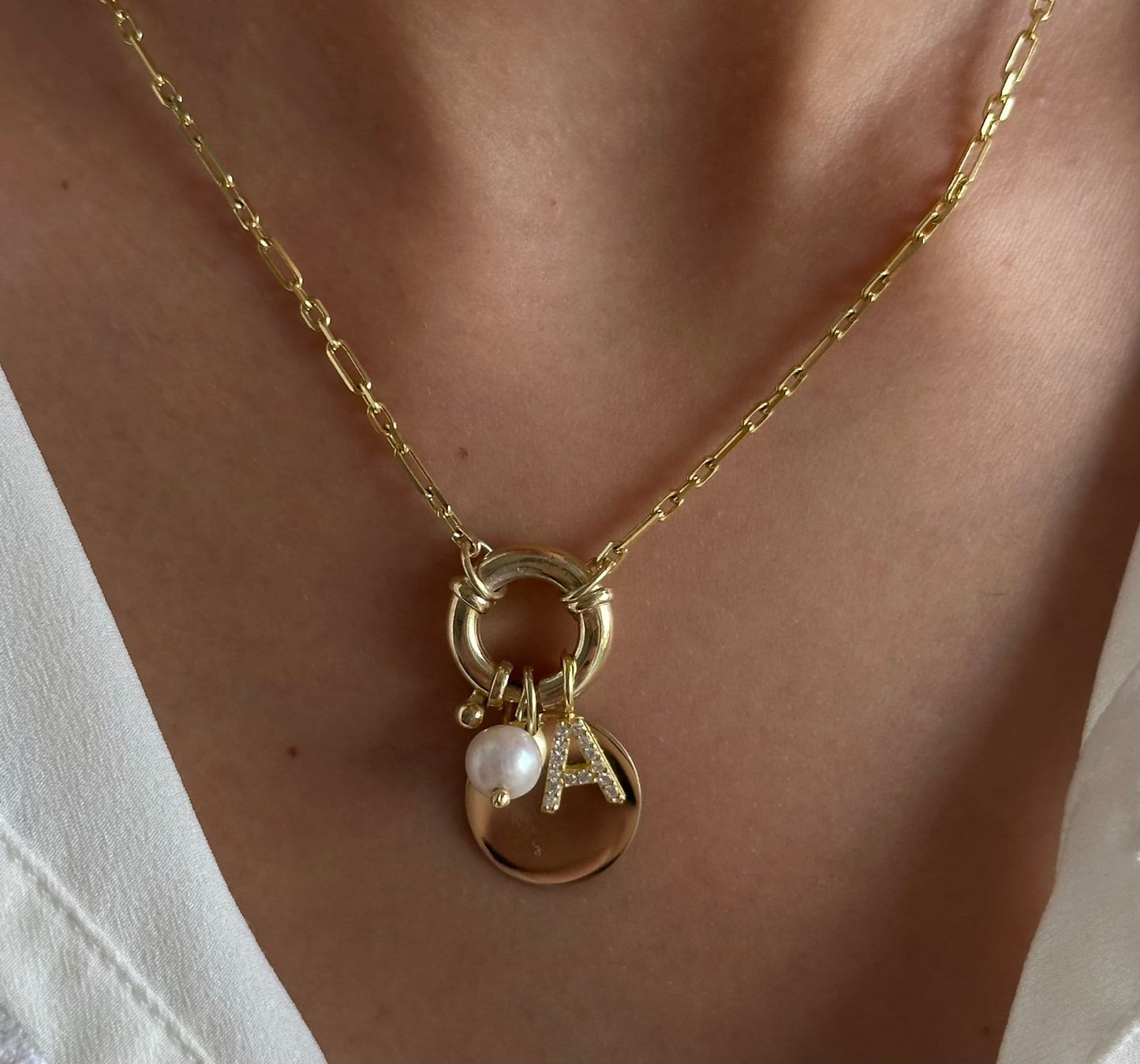 A gold chain necklace with a charm featuring a pearl, a small gold bead, and a gold disc with the letter "A" in sparkling rhinestones.