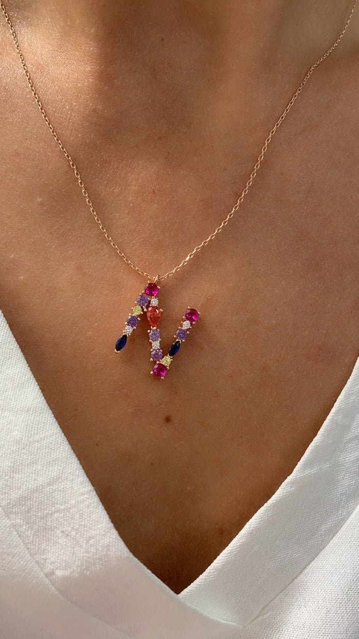 May include: A gold chain necklace with a pendant in the shape of the letter N. The letter is decorated with colorful gemstones, including pink, red, blue, purple, and yellow.
