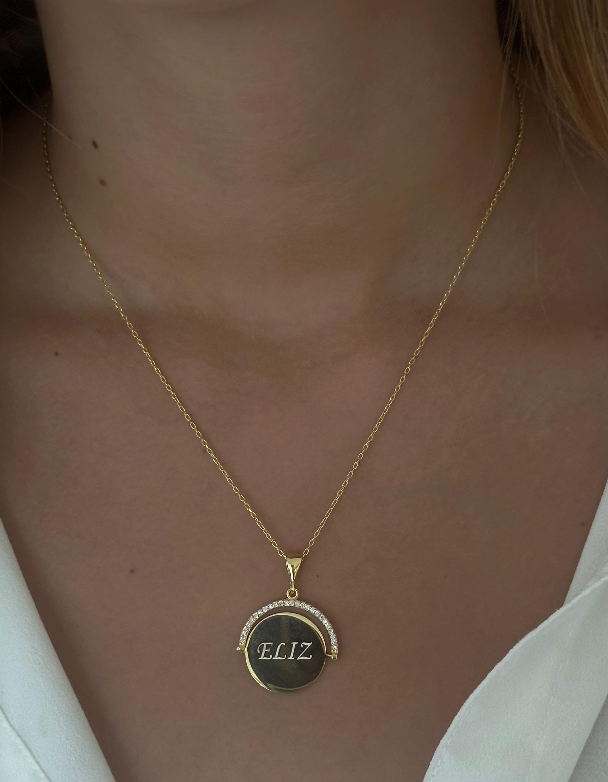 May include: Gold necklace with a spinning pendant. The pendant is round and features the name 'ELIZ' engraved on it.  The pendant's edge is adorned with small, clear cubic zirconia stones. The necklace features a delicate gold chain.