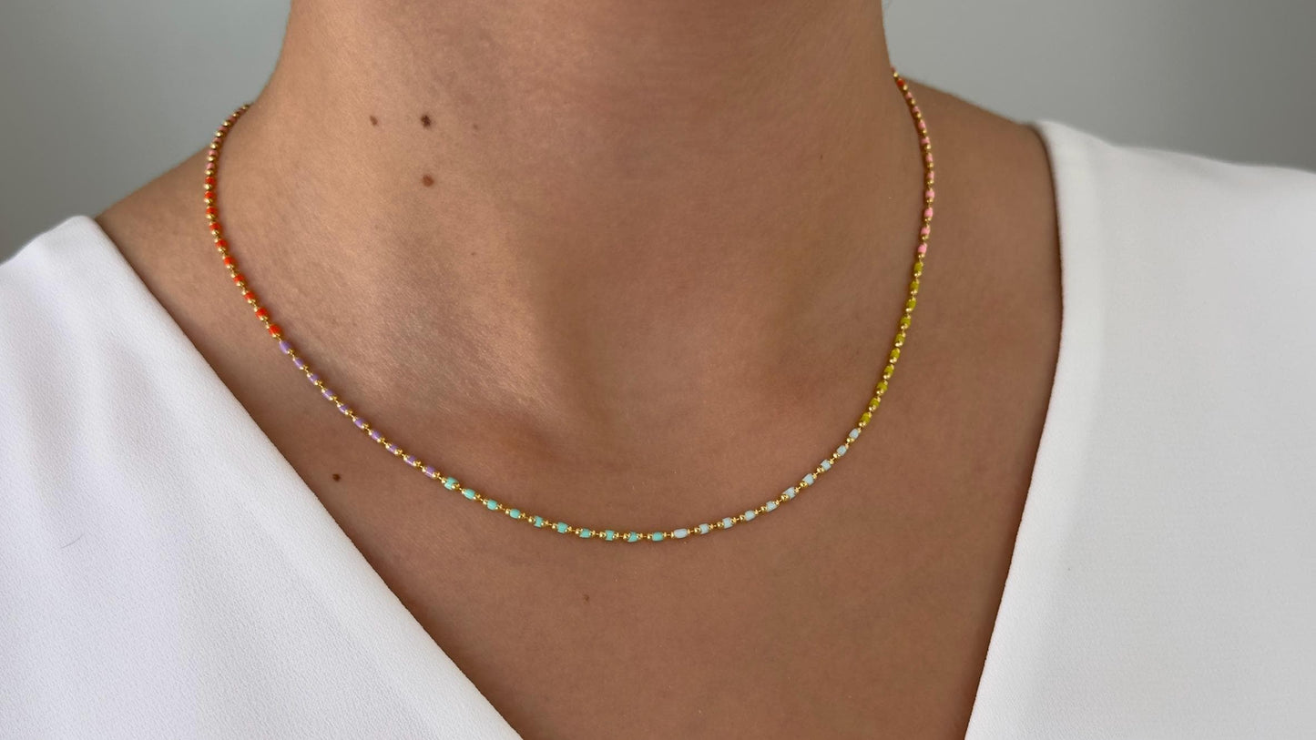 May include: A delicate gold necklace with small, colorful beads. The beads are arranged in a gradient pattern, starting with orange and transitioning to yellow, green, and white.