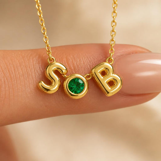 Close-up of a personalized gold birthstone initial necklace featuring polished letter charms and a bezel-set gemstone, shown on a fingertip to highlight the dainty size and premium craftsmanship.