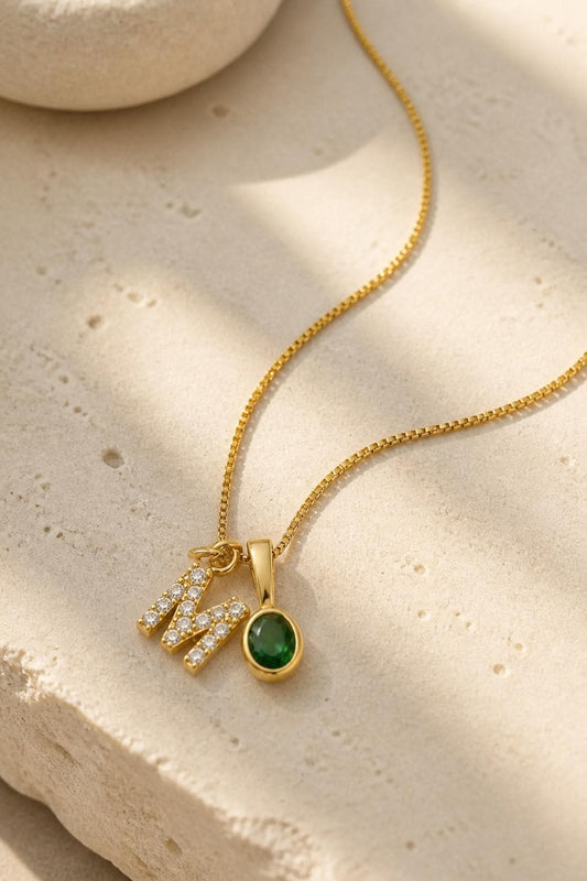 Dainty gold initial necklace with pavé M letter and emerald green oval birthstone charm displayed on stone