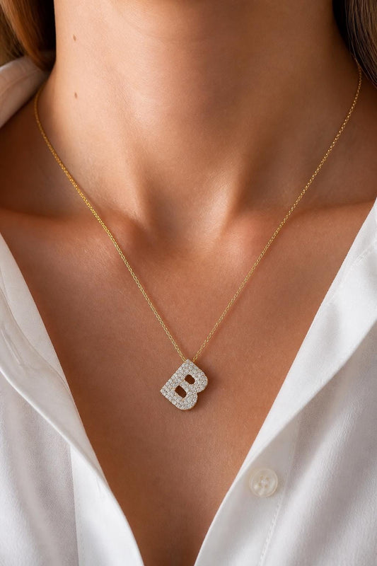 May include: A gold chain necklace featuring a letter B pendant. The pendant is encrusted with small, clear, sparkling stones that cover the entire surface of the letter. The necklace is worn around the neck, resting against a white collared shirt.