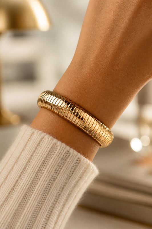 May include: A gold-toned bracelet featuring a segmented, flexible snake-like chain design with a polished, reflective finish. The bracelet wraps around the wrist, showcasing a series of interconnected, rounded metal links that create a smooth, uniform texture.